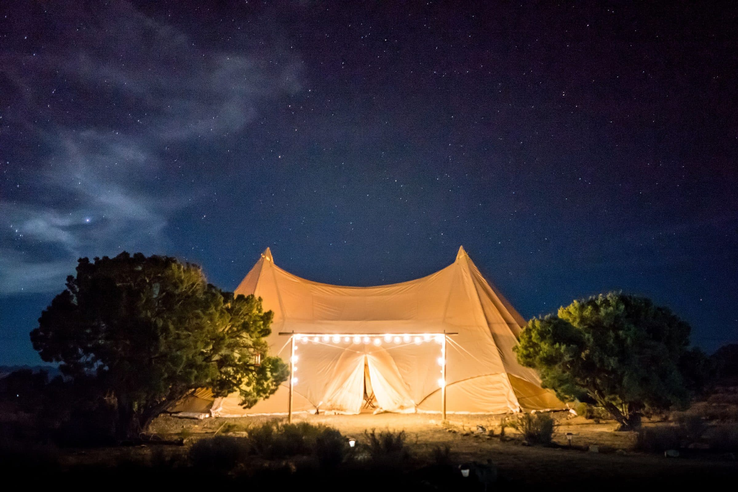 A canvas glamping tent lit with string lights under a starry desert sky.